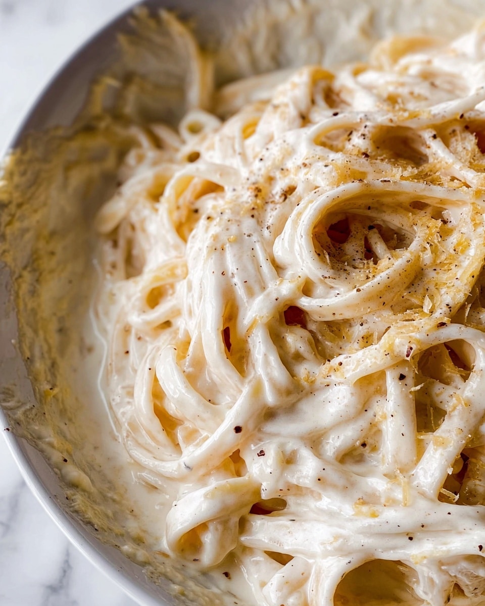 The image shows a close-up of creamy white fettuccine pasta coated in a thick, smooth, white Alfredo sauce. The noodles are twisted and layered unevenly, covered with small specks of black pepper and sprinkled with light yellowish grated cheese. The pasta sits in a white bowl with some sauce clinging to the sides, and the whole scene is set against a white marbled surface. The textures highlight the creaminess of the sauce and the soft, slightly glossy noodles. Photo taken with an iphone --ar 4:5 --v 7