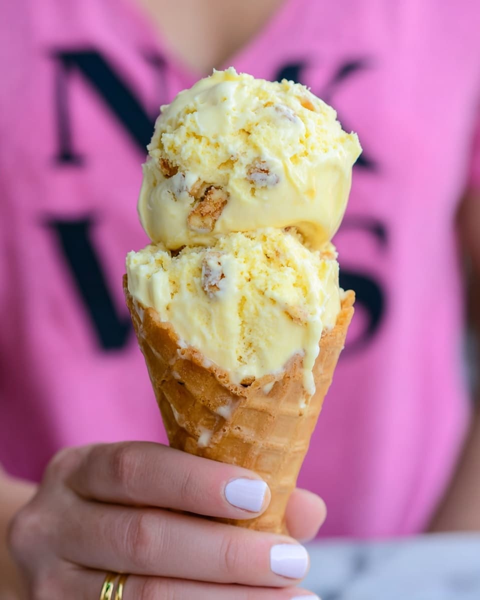 A close-up image of two scoops of creamy yellow ice cream with bits of cake or cookie mixed inside, placed in a light golden waffle cone. The ice cream looks soft and slightly melted, with smooth, glossy drips running down the cone. The cone is held delicately by a woman's hand with French manicure and a thin gold ring on one finger. The background is blurred, showing a person wearing a pink shirt with black block letters. The photo is set on a white marbled surface. photo taken with an iphone --ar 4:5 --v 7
