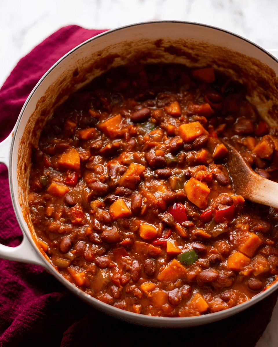 A thick stew with a chunky texture, filled with layers of brown beans, orange sweet potato pieces, and small bits of red and green peppers, all mixed in a rich, reddish-brown sauce. The stew sits inside a white pot with a wooden spoon partially visible on the right side. The inside edge of the pot shows some sauce stains, hinting at slow cooking. The pot rests on a wrinkled dark red cloth on a white marbled surface. Photo taken with an iphone --ar 4:5 --v 7