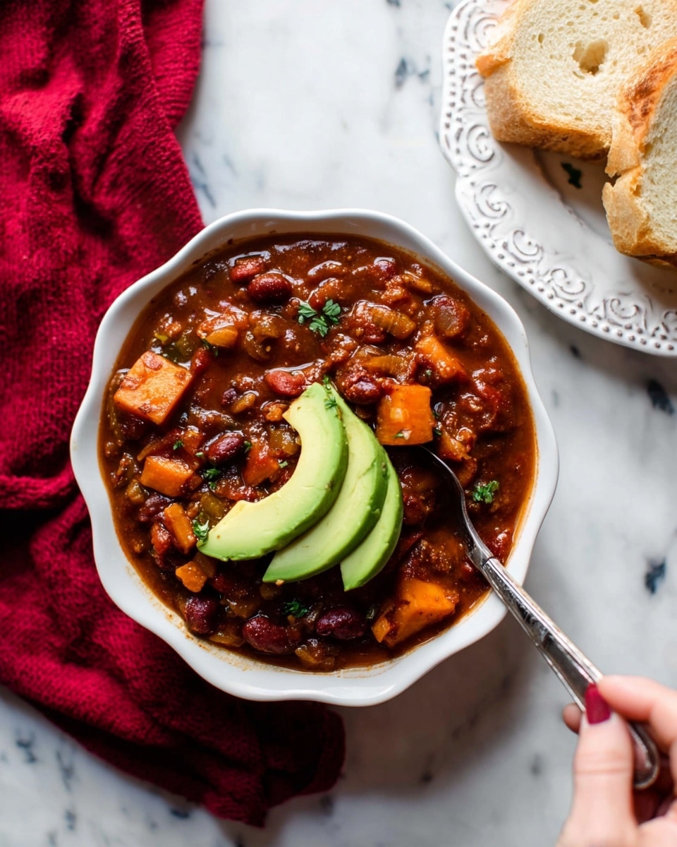 A bowl of thick chili with a deep reddish-brown sauce is shown with visible pieces of beans, diced vegetables, and chunks of sweet potato, creating a chunky texture. On top of the chili, there are two slices of fresh light green avocado placed neatly in the center. The chili fills a white bowl with scalloped edges, set on a smooth white marbled surface. To the top right, some slices of light brown bread rest on a white plate with decorative embossed patterns. A woman's hand on the right side holds a spoon digging into the chili. In the background, a red cloth adds a warm contrast. Photo taken with an iphone --ar 4:5 --v 7