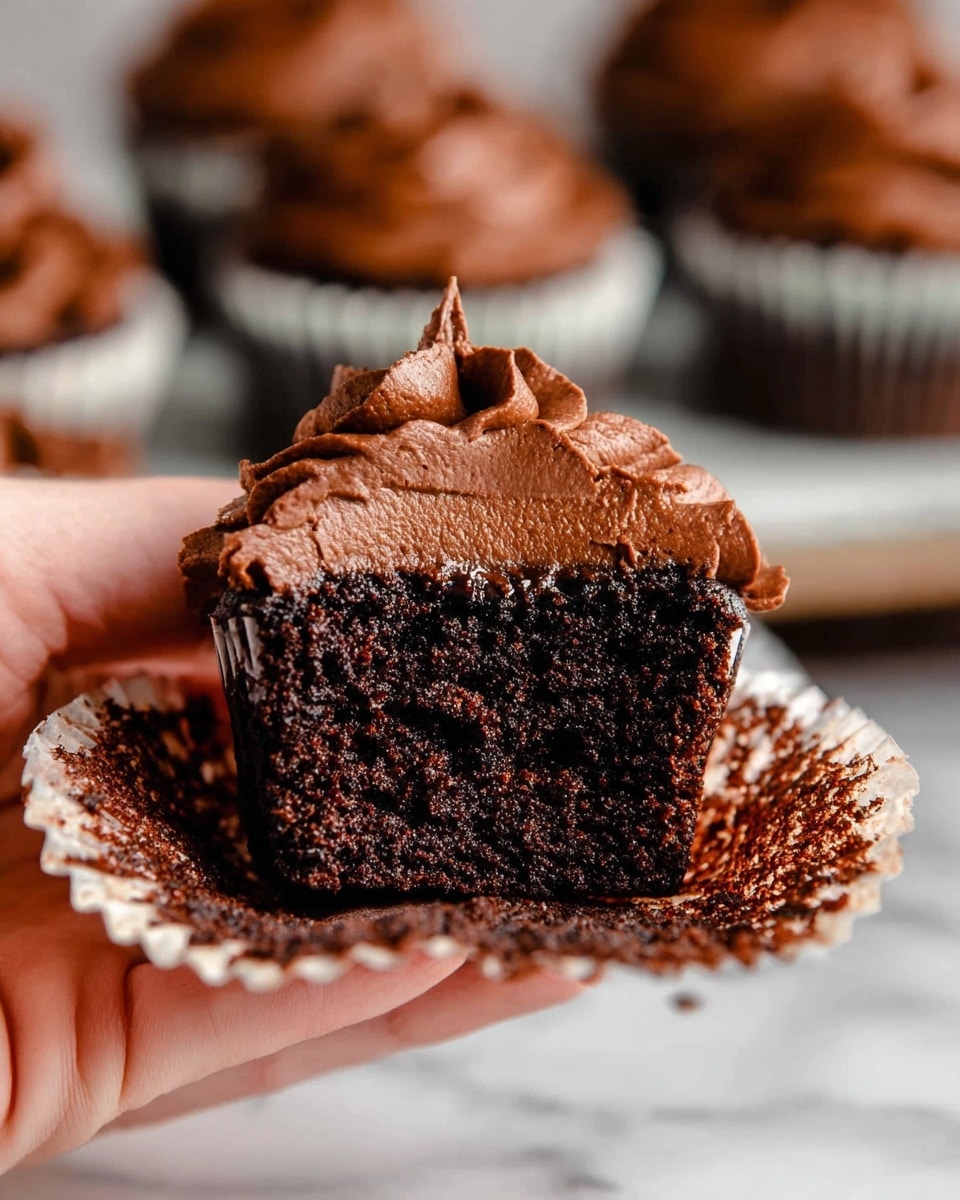 A close-up of a half-eaten chocolate cupcake held by a woman's hand, showing two layers: the bottom layer is dark, moist chocolate cake with a dense texture, and the top layer is smooth, thick chocolate frosting piped in a ruffled swirl. The cupcake liner, peeled back and resting below, is stained dark brown from the cake. In the background, blurred, are more frosted chocolate cupcakes inside white cupcake liners, all set on a white marbled surface. photo taken with an iphone --ar 4:5 --v 7