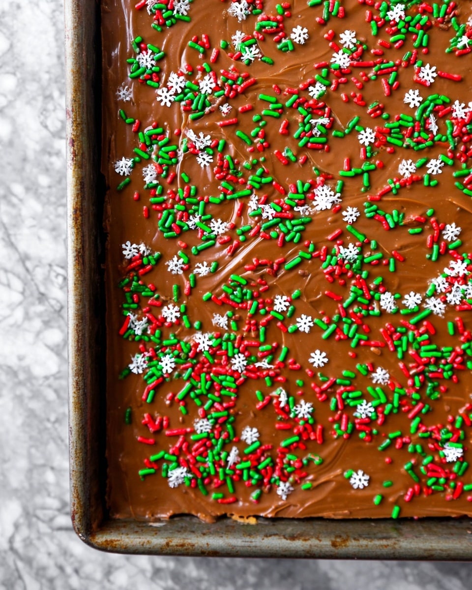 A rectangular dessert bar with one visible layer topped with a thick, smooth layer of shiny milk chocolate that has small swirls and ridges. The chocolate layer is covered evenly with tiny cylindrical red and green sprinkles and small white flower-shaped sprinkles scattered across the top. The dessert sits inside a metal baking pan with slightly worn edges, placed on a surface with a white marbled texture. Photo taken with an iphone --ar 4:5 --v 7