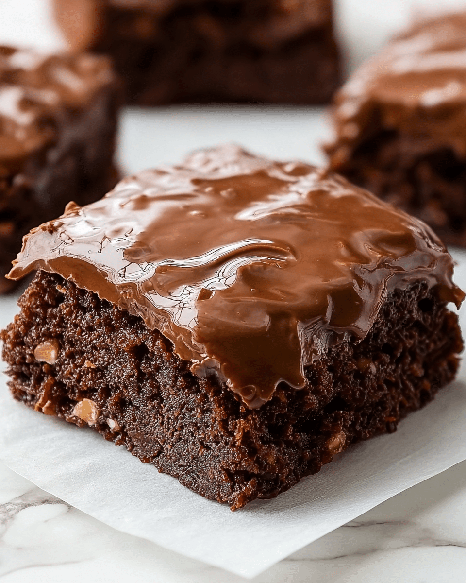 A close-up of a single square piece of chocolate brownie with two other pieces blurred in the background, all on white parchment paper over a white marbled surface. The brownie has two visible layers: a thick, dark brown, dense cake base with a slightly crumbly texture, and a thick, shiny, smooth chocolate glaze on top that appears slightly cracked and glossy, with small nut-like pieces embedded within the glaze. The glaze gently spills over the edges onto the parchment paper. photo taken with an iphone --ar 4:5 --v 7