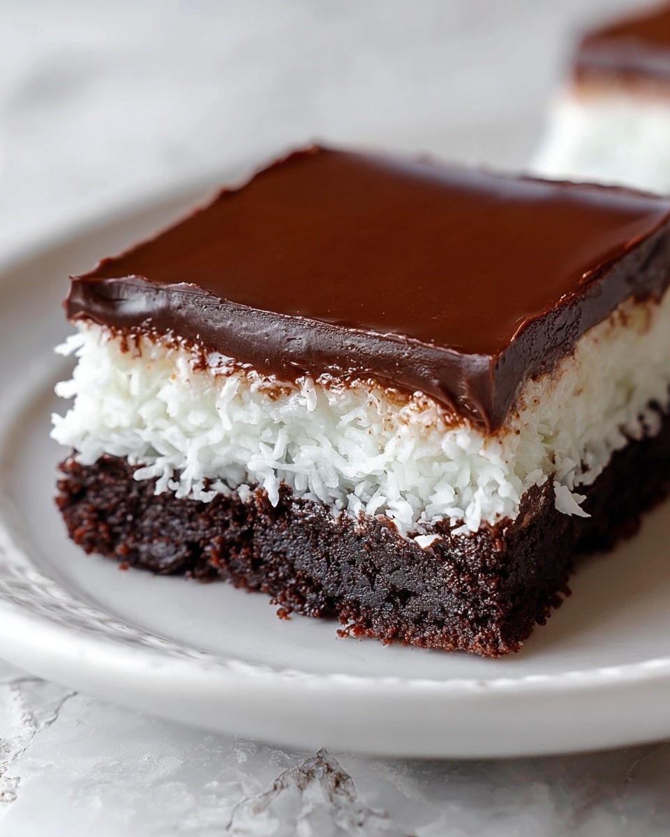 A square dessert with three clear layers sits on a white plate with a subtle pattern. The bottom layer is dark brown, crumbly, and looks like a dense chocolate cake or brownie. Above it is a thick, white layer with a slightly rough texture, resembling shredded coconut or a moist coconut filling. The top layer is a smooth, glossy dark chocolate ganache that evenly covers the dessert. The background is a white marbled texture, and the overall lighting softens the rich colors of the dessert. photo taken with an iphone --ar 4:5 --v 7