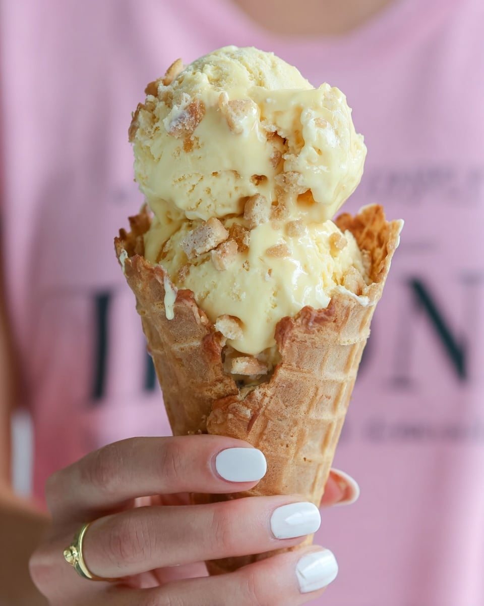 A close-up of a vanilla ice cream cone held by a woman's hand with a thin gold ring and French manicure nails. The cone is light brown with a waffle texture, filled with two scoops of pale yellow ice cream mixed with small cookie or cake chunks. Creamy sauce drips over the ice cream, creating a soft, glossy look. The background is a blurred pink shirt with black text. The image sits on a white marbled texture. photo taken with an iphone --ar 4:5 --v 7