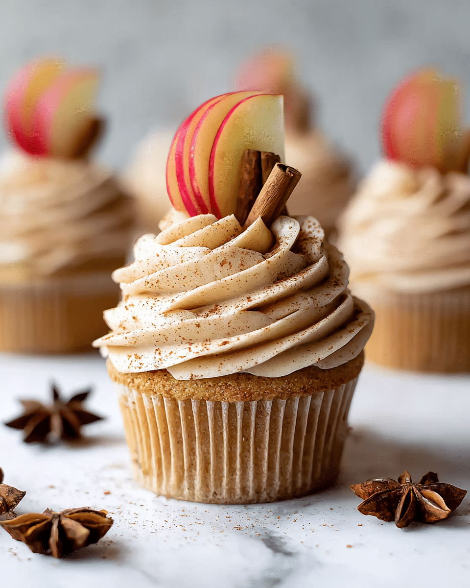 A single cupcake sits on a white marbled surface with a golden brown base that looks soft and fluffy. On top, there is a thick, swirled layer of creamy, light beige frosting dusted lightly with cinnamon powder. Stuck into the frosting is a thin slice of red and yellow apple, standing upright, with two small cinnamon sticks placed behind the apple slice. Around the cupcake, there are star anise pieces scattered on the white marbled surface. Several similar cupcakes appear blurred in the background, all decorated the same way. photo taken with an iphone --ar 4:5 --v 7