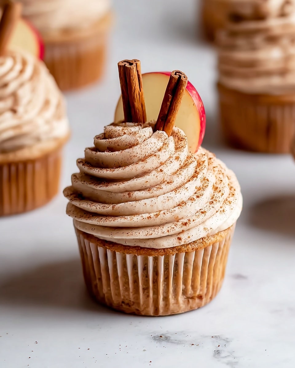 The image shows a close-up of a cupcake with three main layers. The base layer is a light brown cupcake with a soft texture. On top, there is a swirl of creamy frosting in a light beige color, sprinkled with a light dusting of brown spice powder. The frosting has a smooth and whipped texture, forming multiple rounded ridges. The topmost layer includes a half slice of red apple placed on one side of the frosting and two vertical cinnamon sticks inserted next to the apple slice. The cupcakes are set on a white marbled surface. photo taken with an iphone --ar 4:5 --v 7
