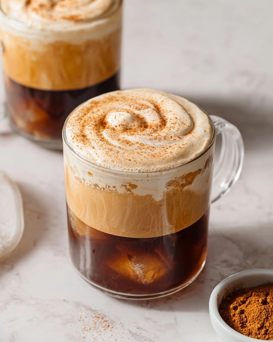The image shows two clear glass mugs filled with a three-layered coffee drink placed on a white marbled surface. The bottom layer is dark brown iced coffee with visible ice cubes. The middle layer is a light brown frothy mix that gradually blends into the coffee below. The top layer is thick, creamy foam with a beige color sprinkled with fine cinnamon powder. One mug is in the front and the other is partially visible in the back, both with smooth, swirled texture on top. To the side, a small white bowl contains loose cinnamon powder. The scene is softly lit, emphasizing the creamy texture and warm colors of the drink. photo taken with an iphone --ar 4:5 --v 7
