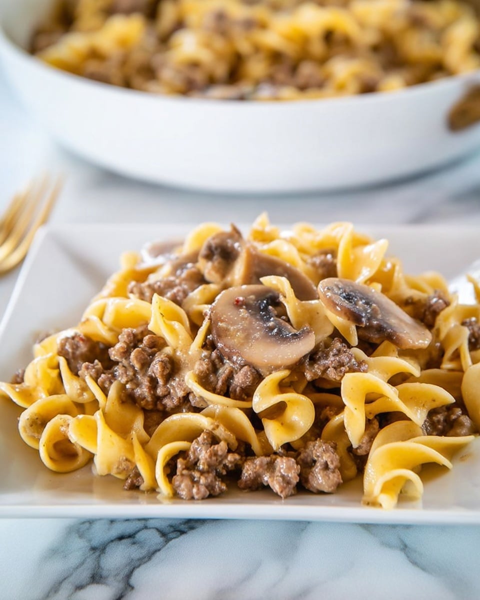 The dish shows a close-up of a white square plate filled with wide, curly buttered noodles mixed generously with browned ground beef crumbles and several slices of cooked brown mushrooms. The noodles have a yellowish color and a soft texture, while the beef looks tender and juicy. The mushrooms add an earthy brown tone, and the sauce lightly coats the ingredients, giving a slight shine. In the blurred background, there is a white bowl with more of the same dish. The surface underneath the plate is white with a marbled texture. photo taken with an iphone --ar 4:5 --v 7