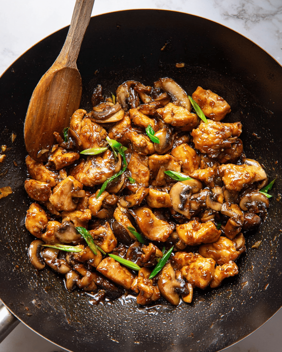 The dish is shown inside a deep black wok, filled with many pieces of golden brown, crispy chicken that have a slightly shiny texture from sauce. Mixed among the chicken are slices of cooked mushrooms with a soft, brown surface and pieces of green onion adding green color. A wooden spoon coated with sauce rests on the left side of the wok. The dish looks juicy and well coated with a thick, dark brown sauce. The background is a white marbled texture photo taken with an iphone --ar 4:5 --v 7