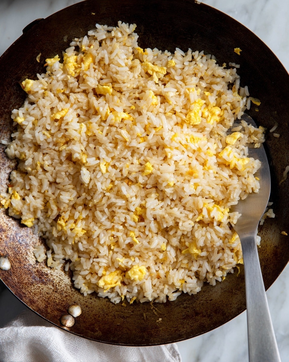 A close-up view of a dark, worn frying pan filled with cooked fried rice, showing white rice grains mixed with small pieces of light yellow scrambled eggs and small bits of onions, creating a textured and slightly oily appearance. The rice looks fluffy and lightly browned in some spots from frying, and a metal spatula is placed partially under the rice in the pan. The pan rests on a white marbled surface with a light cloth napkin partially visible in the background. photo taken with an iphone --ar 4:5 --v 7
