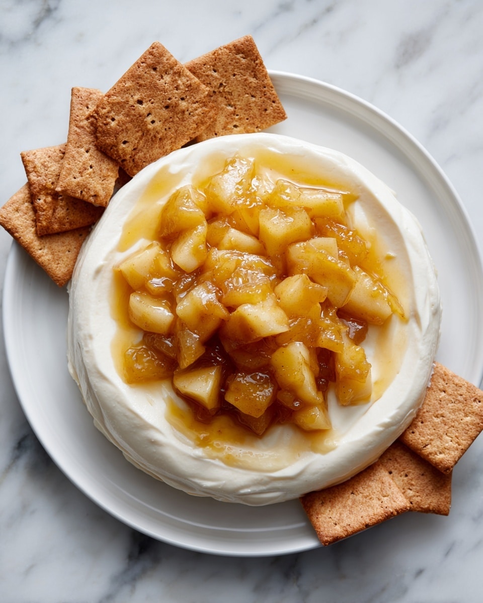 A white plate holds a two-layer dessert placed on a white marbled surface. The bottom layer is smooth and creamy, off-white in color, spread in a ring shape with a thick center left open. On top, there is a glossy, chunky layer of cooked apple pieces in a warm golden-brown syrup, filling the center of the cream. A rectangular brown cracker is partially stuck into the dessert on one side, with several more crackers scattered around the plate on the surface. The textures combine soft cream, juicy apple chunks, and crunchy crackers. Photo taken with an iphone --ar 4:5 --v 7