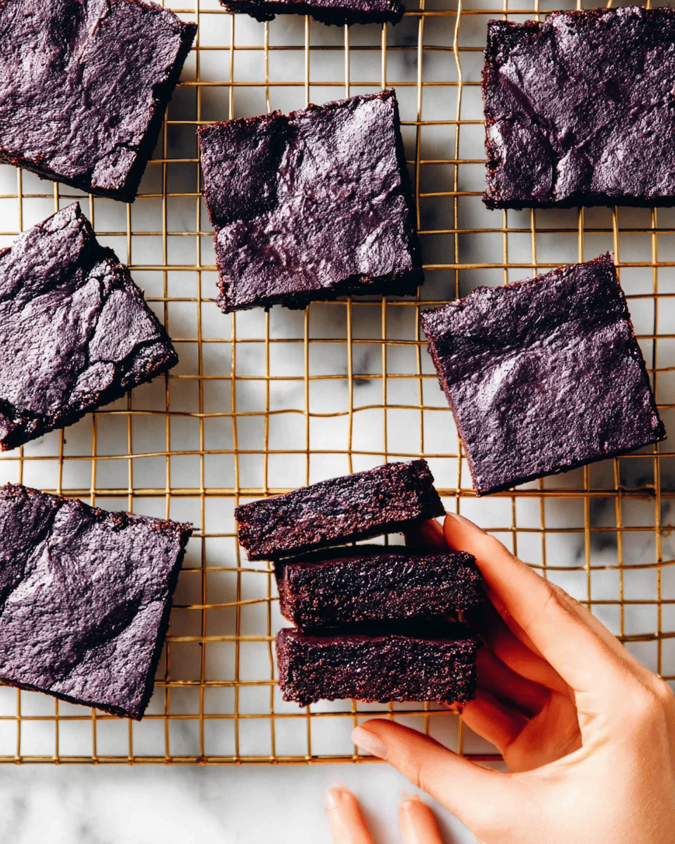 The image shows several square-shaped, dark purple brownies placed on a gold wire cooling rack. Most brownies lie flat, showing a slightly wrinkled, cracked top texture with a deep purple color. One woman's hand at the bottom right is holding a stack of three brownies, revealing a dense, moist inside that is almost black with a thin purple crust on the edges. The background is a white marbled surface, with another woman's hand reaching toward the lower part of the frame. photo taken with an iphone --ar 4:5 --v 7