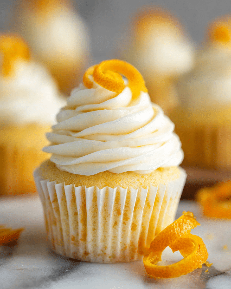 The image shows a close-up of a cupcake with three thick layers of white cream swirled neatly on top. The cupcake base is golden yellow and wrapped in a white paper liner with subtle vertical ridges. There is a small, curly orange peel garnish placed on the peak of the cream, with a few more orange peel curls placed beside the cupcake on a white marbled surface. The background is softly blurred, showing more similar cupcakes. photo taken with an iphone --ar 4:5 --v 7