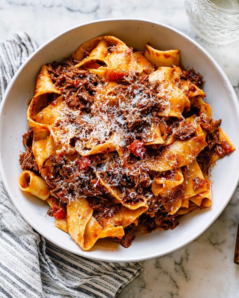 A white bowl filled with thick, flat pasta ribbons layered with a rich, dark red-brown meat sauce that has shredded beef mixed throughout, showing a mix of soft and tender textures. On top, a sprinkling of finely grated white cheese adds a light, snowy contrast to the warm colors of the sauce and pasta beneath. The bowl sits on a white marbled textured surface with a striped cloth napkin and a gold fork nearby, creating a cozy and inviting setting. photo taken with an iphone --ar 4:5 --v 7