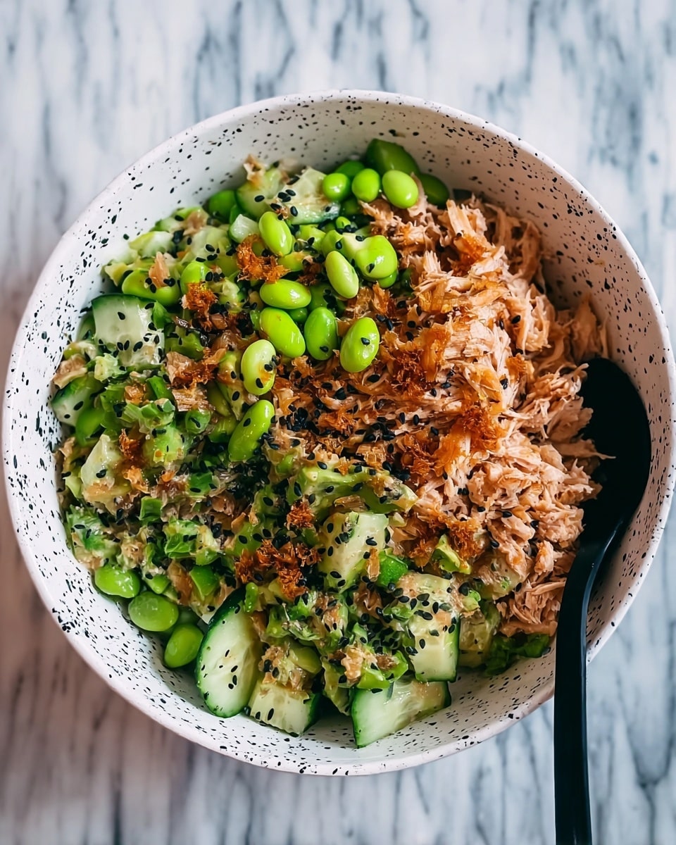 This image shows a bowl of mixed food with a close view. It has several layers: the bottom layer looks like shredded cooked chicken that is light brown and soft. On top, there are many slices of bright green cucumber, some whole green edamame beans, and bits of avocado. There are small pieces of light brown fried onions or garlic spread across, adding a crunchy texture. Black sesame seeds are sprinkled over everything, adding tiny dark spots. The bowl is white with black speckles and the background shows a white marbled texture. A black spoon is placed inside the bowl on the right side. photo taken with an iphone --ar 4:5 --v 7