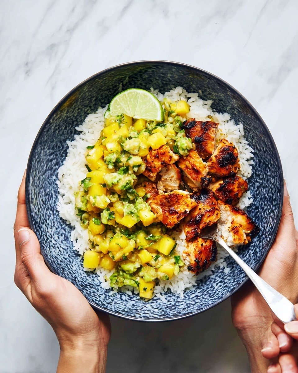 A blue patterned bowl filled with three main layers: white fluffy rice at the base, topped with golden-brown grilled chicken pieces that have a slightly charred texture, and a chunky yellow mango and green avocado salsa scattered over the chicken and rice. There is a wedge of lime tucked slightly behind the chicken on one side. A silver spoon is scooping a piece of chicken from the bowl, which is held by two woman's hands over a white marbled surface. photo taken with an iphone --ar 4:5 --v 7