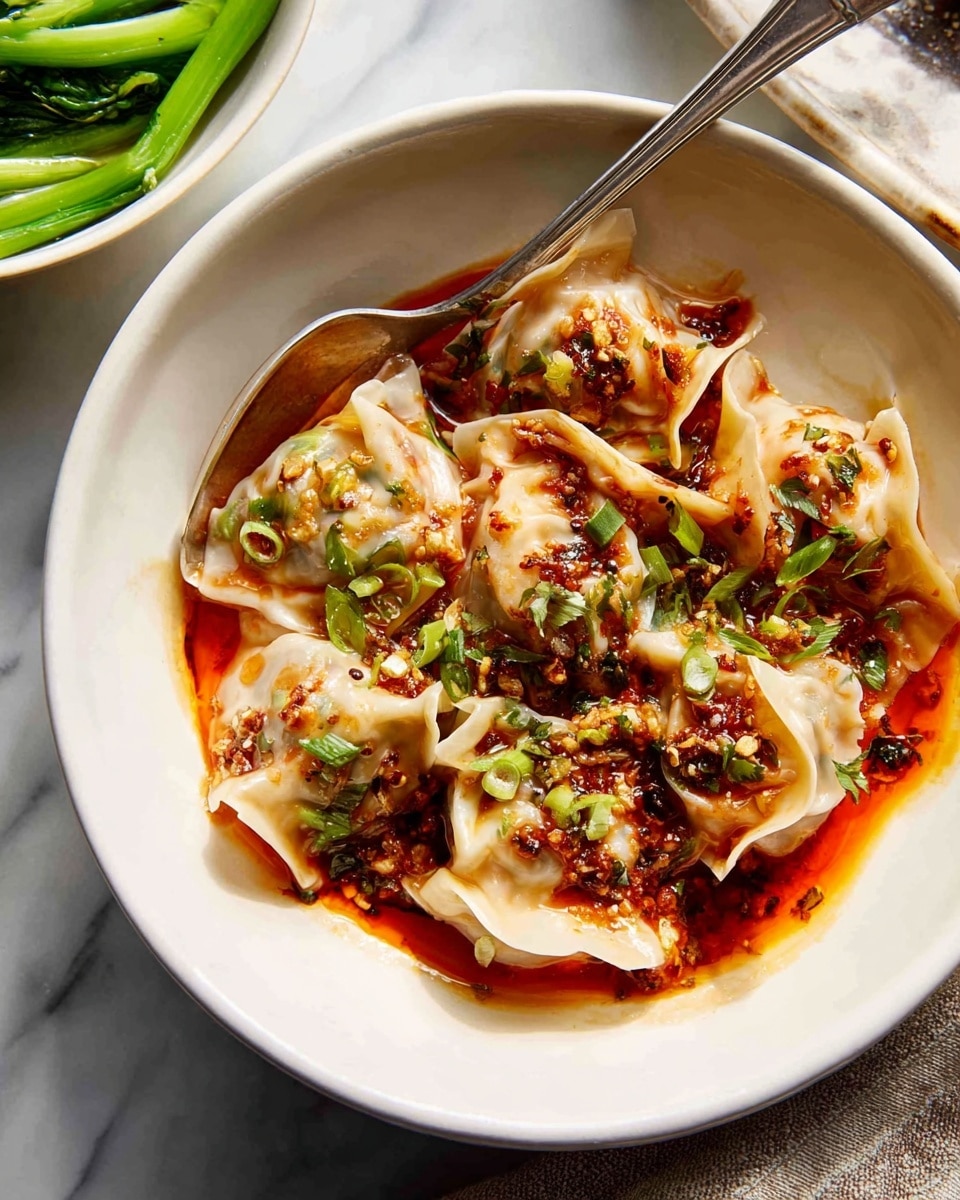 The image shows a white bowl filled with several dumplings covered in a shiny, reddish-brown sauce with bits of green herbs and small pieces of garlic or onion sprinkled on top. The dumplings have a soft, translucent wrapper that is folded and slightly wrinkled. The sauce pools around the dumplings at the bottom of the bowl, adding a rich, glossy texture. A silver spoon rests inside the bowl, partially covered by the sauce and dumplings. In the background, there is a white bowl with green steamed vegetables, sitting on a white marbled surface. photo taken with an iphone --ar 4:5 --v 7