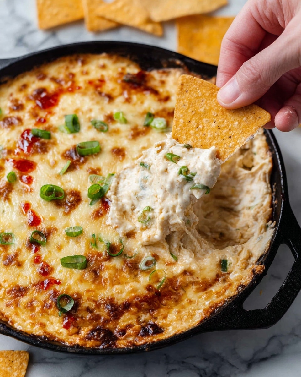 A close-up of a black cast iron skillet filled with a creamy, bubbly cheese dip that has a golden brown, slightly toasted top layer with green sliced scallions sprinkled on it and small drops of vibrant red sauce for color. A woman's hand is dipping a triangle-shaped, golden brown, textured chip into the thick, soft cheese inside the skillet near the bottom right edge. The background surface is a white marbled texture with a few extra chips scattered around. Photo taken with an iphone --ar 4:5 --v 7