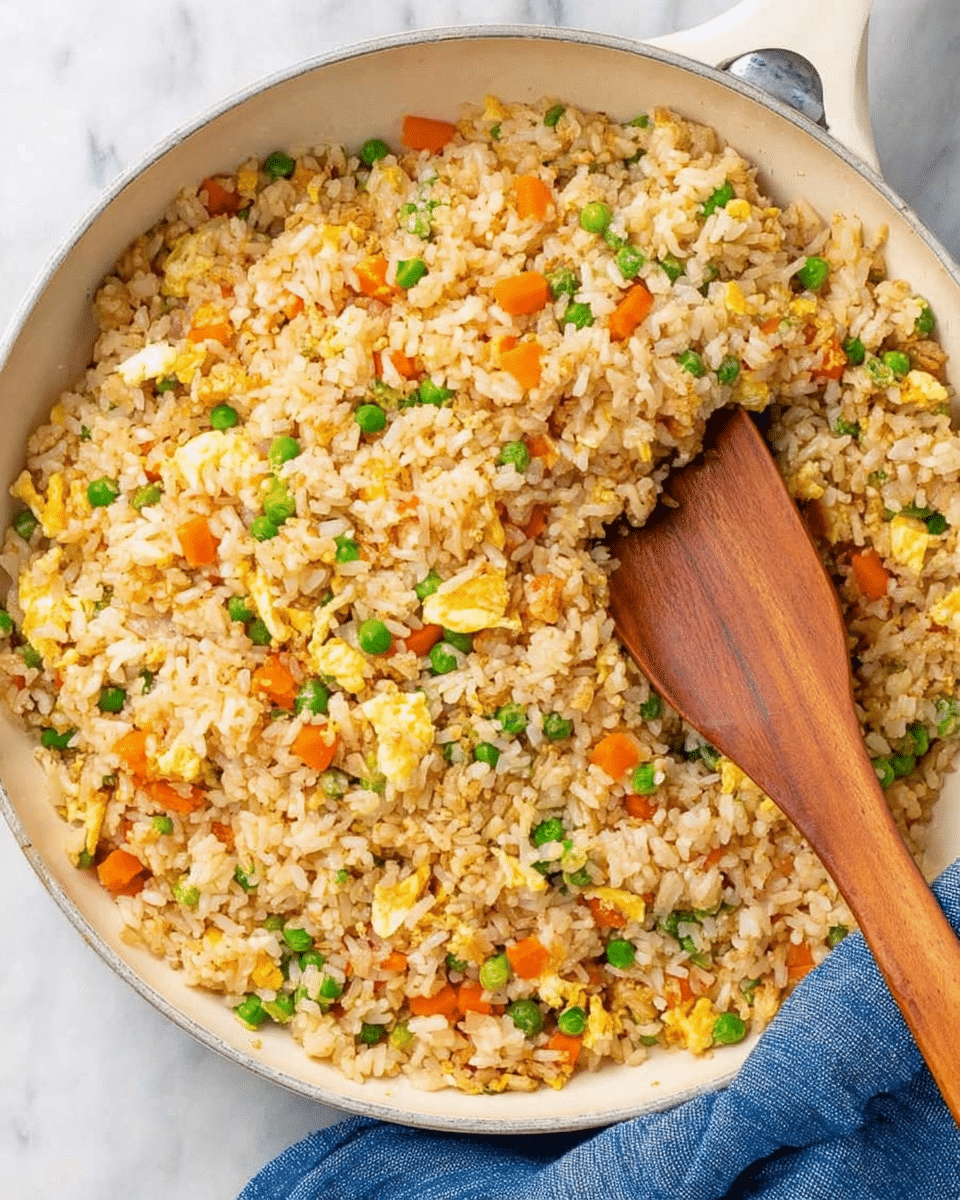 A white skillet filled with fried rice, showing one layer made up of light brown rice grains mixed evenly with small pieces of orange carrots and green peas, along with bits of scrambled egg scattered throughout. On top of the rice is a wooden spatula resting inside, its smooth light brown color contrasting with the textured rice. The skillet sits on a white marbled surface and a blue cloth is partially underneath it. photo taken with an iphone --ar 4:5 --v 7