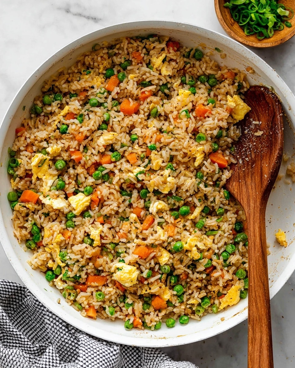 A white skillet filled with fried rice that has about three layers: the main layer of light brown cooked rice mixed with small, bright green peas, diced orange carrots, and dark green chopped scallions, with scrambled eggs in small yellow and white chunks scattered throughout, sprinkled with black pepper. A wooden spoon is partially submerged on the right side, mixing the rice. The skillet sits on a white marbled surface with a gray and white checkered cloth underneath one corner. A small wooden bowl with extra green scallions is partly visible at the top right corner. photo taken with an iphone --ar 4:5 --v 7