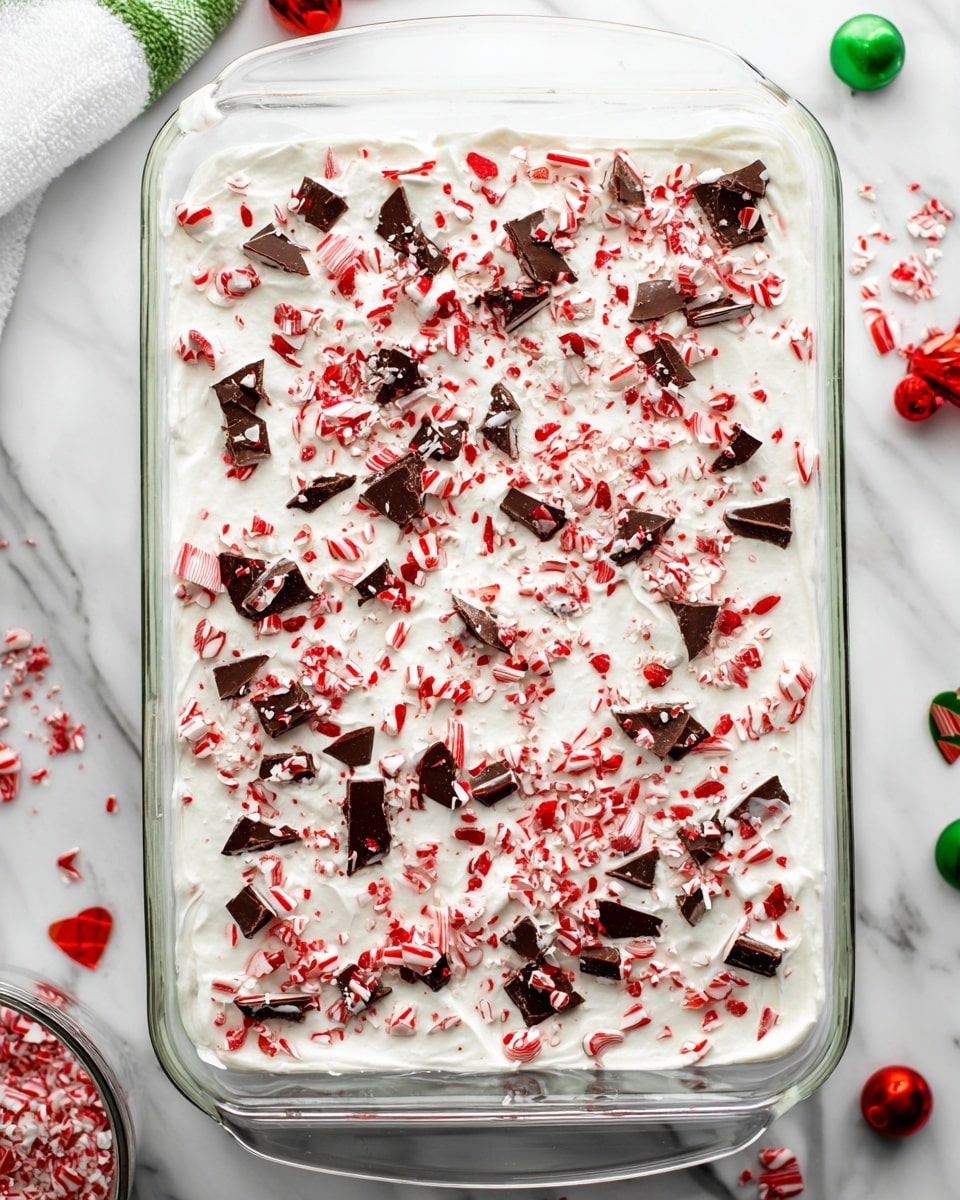 A glass rectangular dish filled with a layered dessert is shown from above. The top layer is smooth and white, likely whipped cream or frosting, spread evenly across the dish. Scattered generously over the top are small broken pieces of dark and red peppermint candies, creating a colorful, slightly rough texture with red, dark brown, and white fragments spread all over. The dish is placed on a white marbled surface with some green and red holiday decorations around it. photo taken with an iphone --ar 4:5 --v 7