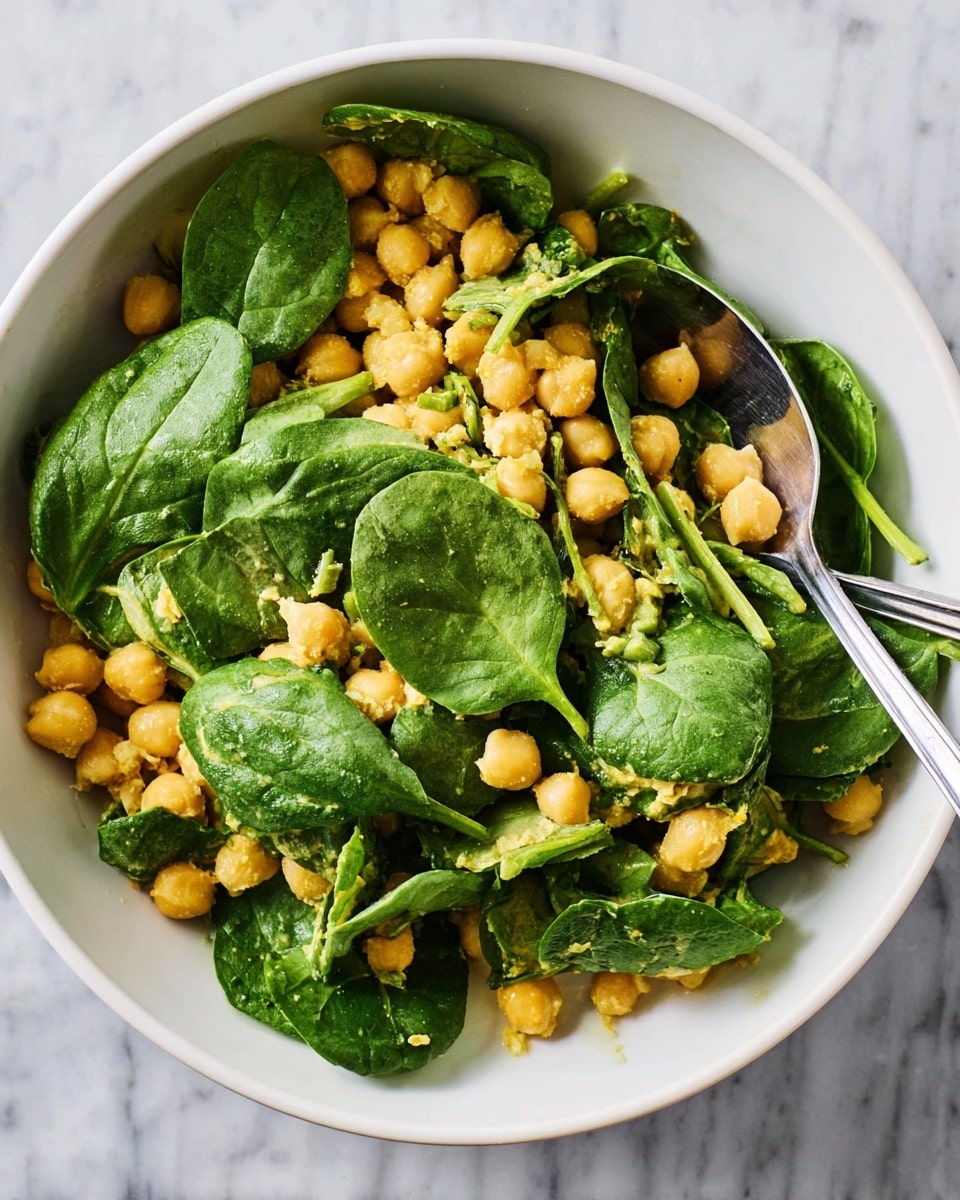 The image shows a white bowl filled with a salad made of two main layers: the bottom layer is light yellow chickpeas with a slightly grainy texture, while the top layer consists of fresh, dark green spinach leaves that are large and have visible veins. Mixed in between the chickpeas and spinach are small pieces of light green chopped herbs or vegetables, adding specks of extra color and texture. A silver fork and spoon rest inside the bowl, partly covered by the salad, and the bowl is set on a white marbled surface. photo taken with an iphone --ar 4:5 --v 7