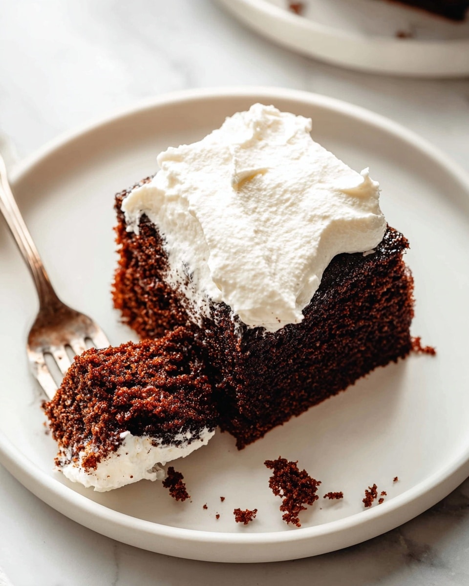 A close-up view of a square piece of dark brown chocolate cake with a rough, slightly crumbly texture, topped with a thick layer of white whipped cream that looks soft and fluffy. The cake sits on a plain white plate placed on a white marbled surface, with a fork holding a bite-sized piece of cake and whipped cream on the left side of the plate. Small crumbs of cake are scattered near the cake and fork, emphasizing its crumbly texture. Photo taken with an iphone --ar 4:5 --v 7