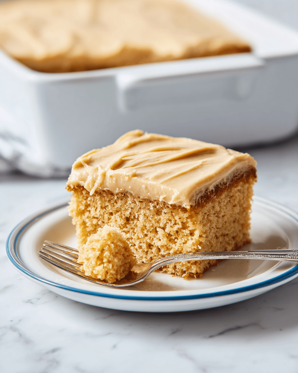 A square piece of moist, light brown cake with a smooth, creamy, tan-colored frosting layer on top, sitting neatly on a white plate with a blue rim. A fork rests on the plate, holding a small chunk of the cake with frosting, showing its soft texture. The background is a white marbled surface with a white, rectangular baking dish slightly blurred in the back. Photo taken with an iphone --ar 4:5 --v 7