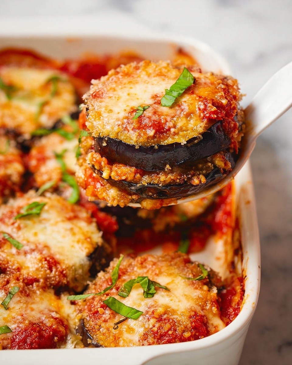 A white dish filled with multiple stacked rounds of eggplant slices layered with red tomato sauce, a thick layer of melted golden cheese on top, and small fresh green basil leaves scattered on each piece. Each stack has at least two visible dark purple eggplant slices with tomato sauce and cheese layers in between. A white spoon is lifting one stack, showing the soft, layered texture. The dish sits on a white marbled surface. photo taken with an iphone --ar 4:5 --v 7