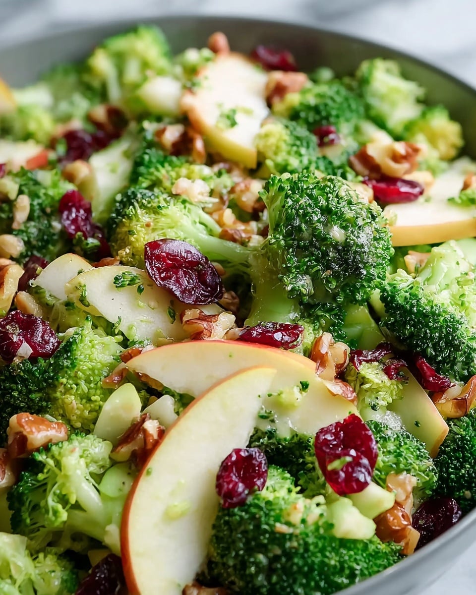 A colorful broccoli salad is shown close up with multiple layers: bright green broccoli florets form the main layer with a fresh, bumpy texture; scattered among the broccoli are thin, pale yellow apple slices with a smooth surface; deep red dried cranberries add small pops of color evenly spread throughout; crunchy, light brown walnut pieces are scattered on top, adding texture contrast; tiny green herb pieces are visible dotting the salad. The salad is on a white marbled surface. photo taken with an iphone --ar 4:5 --v 7