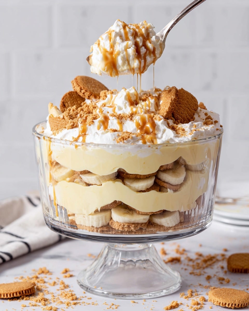 A clear glass trifle bowl with a pedestal holds a layered dessert with four main layers: the bottom layer is a creamy pale yellow pudding, followed by a layer of round golden biscuits and sliced bananas, then another thick layer of the same pale pudding, topped with more biscuits and banana slices, a generous fluffy white whipped topping, and a final drizzle of light brown caramel or butterscotch sauce along with crushed biscuit crumbs on top. A spoon is lifting a spoonful from the bowl showing the soft, creamy, and crunchy textures mixed together, with the background being a white marbled texture and some scattered biscuit crumbs around. photo taken with an iphone --ar 4:5 --v 7