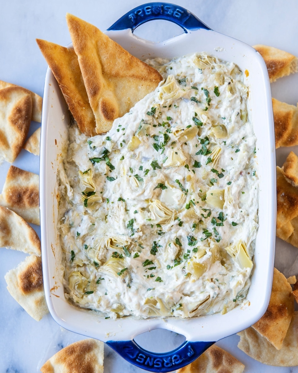 A white rectangular baking dish with blue handles is filled with a creamy dip that has a soft, thick texture. The dip is mainly white with small green herbs sprinkled evenly on top, giving a fresh look. There are visible pieces of pale yellow artichoke hearts mixed inside the dip. Three golden-brown pita chips are placed upright, partly dipped into the creamy mixture at the corner of the dish. The dish is set on a white marbled surface with some extra pita chips scattered around. photo taken with an iphone --ar 4:5 --v 7