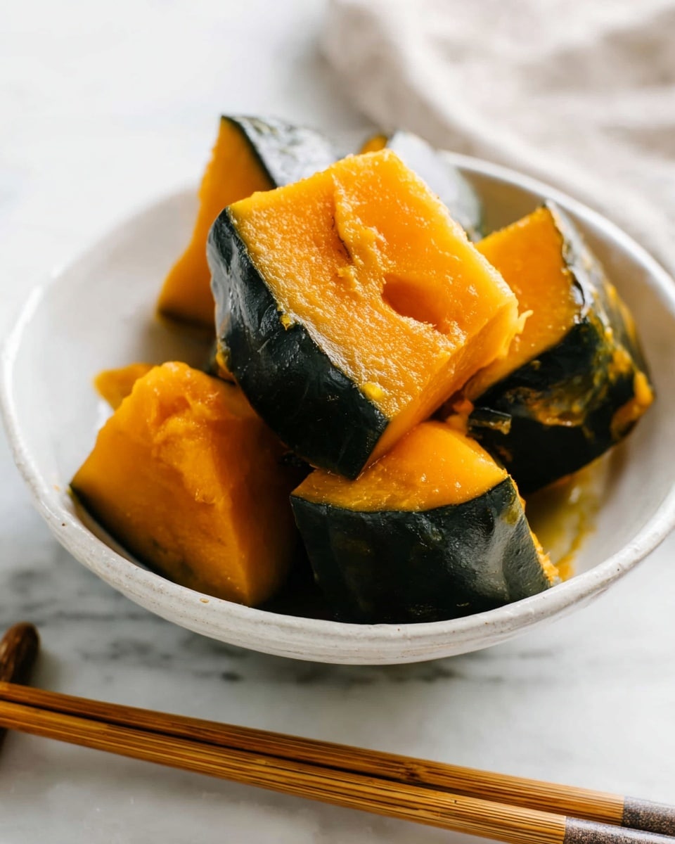 A white bowl filled with several thick chunks of cooked pumpkin, each piece showing a bright orange interior with dark green skin edges. The pumpkin pieces have a soft, slightly glossy texture and are stacked loosely inside the bowl. Next to the bowl on a white marbled surface are a pair of wooden chopsticks with natural grain and color visible. The setting is bright and clean, highlighting the rich colors of the pumpkin. photo taken with an iphone --ar 4:5 --v 7