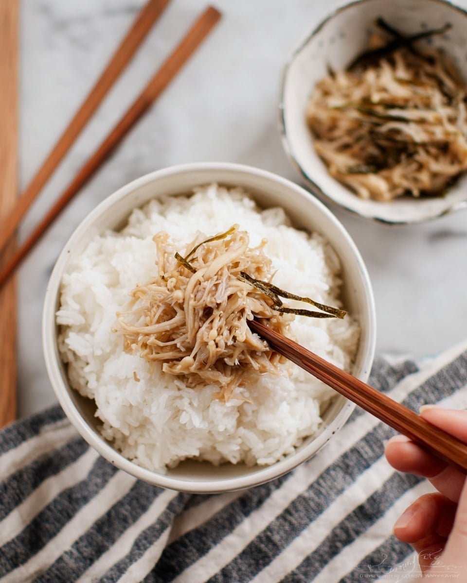 A white bowl filled with a thick layer of fluffy, white cooked rice serves as the base. On top, light brown enoki mushrooms with thin, stringy textures and a few greenish seaweed strips are being picked by wooden chopsticks held by a woman's hand from the upper right corner. Nearby, a small white bowl contains more of the mushroom topping, and the scene is set on a white marbled textured surface with a striped cloth partially visible under the bowls. photo taken with an iphone --ar 4:5 --v 7