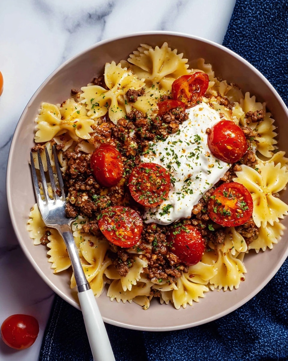 A white bowl is filled with one layer of cooked bowtie pasta, pale yellow and soft. On top of the pasta is a large dollop of white creamy sauce, smooth in texture. Above this is a generous layer of browned minced meat mixed with small bits of cooked onion, dark brown with a crumbly look. Scattered on top are several bright red cherry tomato halves, glossy and fresh, sprinkled with green dried herbs. A silver fork with a white handle rests on the left side of the bowl. The bowl sits on a white marbled surface with a navy blue cloth partly visible at the bottom edge. Photo taken with an iphone --ar 4:5 --v 7