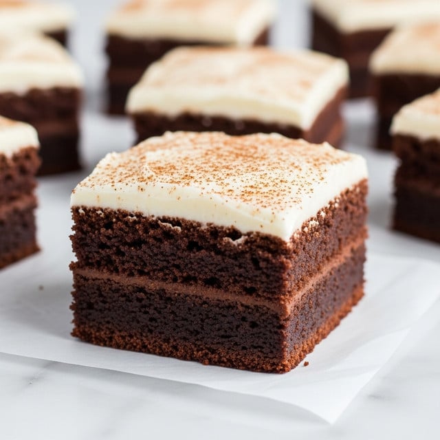A close-up image of a square chocolate brownie with two clear layers: the bottom layer is dark, rich, and moist chocolate cake with a slightly crumbly texture, while the top layer is a thick, creamy white frosting dusted lightly with brown spice powder. The brownie is placed on a sheet of white parchment, resting on a white marbled surface, with several other similarly frosted brownie squares blurred softly in the background. photo taken with an iphone --ar 4:5 --v 7
