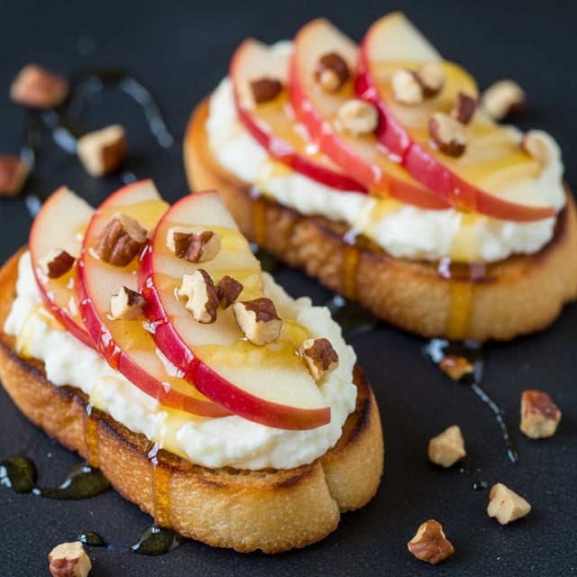 Two small slices of toasted bread serve as the base for this dish, each topped with a layer of melted creamy cheese that is smooth and white. Above the cheese, thin slices of red-skinned apple are neatly placed, showcasing a light yellow interior with the red skin visible on the edges. Small chunks of toasted brown nuts are scattered on top of the apples, adding texture and color contrast. A light golden honey drizzle glistens over the nuts, apples, and cheese, creating a shiny finish. The toasts are placed on a dark surface with scattered nut pieces around them. Photo taken with an iphone --ar 4:5 --v 7