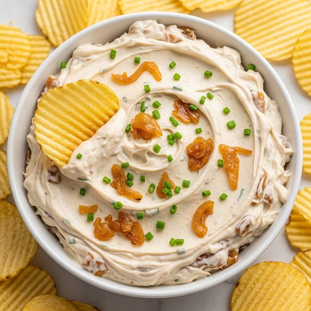 A close-up view of a bowl filled with creamy white dip mixed with visible caramelized light brown onion pieces, topped with small green chopped chives. The dip has a thick, textured swirl pattern on the surface, and two ridged yellowish potato chips are dipped into it on the left side. The bowl is white, round, and shallow, placed on a smooth white marbled surface, surrounded by more potato chips scattered around it. photo taken with an iphone --ar 4:5 --v 7