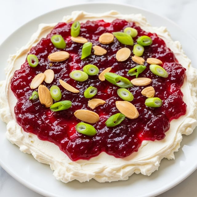 A close-up view of a creamy white base layer with a smooth, thick texture on a white plate, topped with a shiny, deep red cranberry sauce that has a chunky, gel-like texture spread unevenly across the surface. Scattered on top are thin, light brown almond slices with a slightly glossy finish, and fresh bright green sliced green onions that add a fresh contrast. The white plate sits on a white marbled surface. photo taken with an iphone --ar 4:5 --v 7