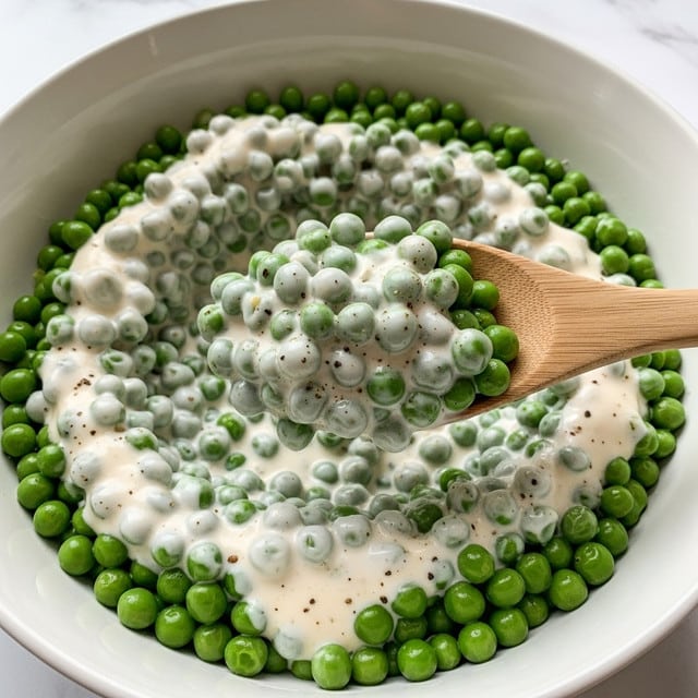 A close-up image shows a large white bowl filled with green peas covered in a thick, creamy white sauce with specks of black pepper. A wooden spoon lifts a portion of the peas, highlighting their bright green color and the smooth, rich texture of the creamy sauce coating each pea evenly. The background has a white marbled texture. photo taken with an iphone --ar 4:5 --v 7