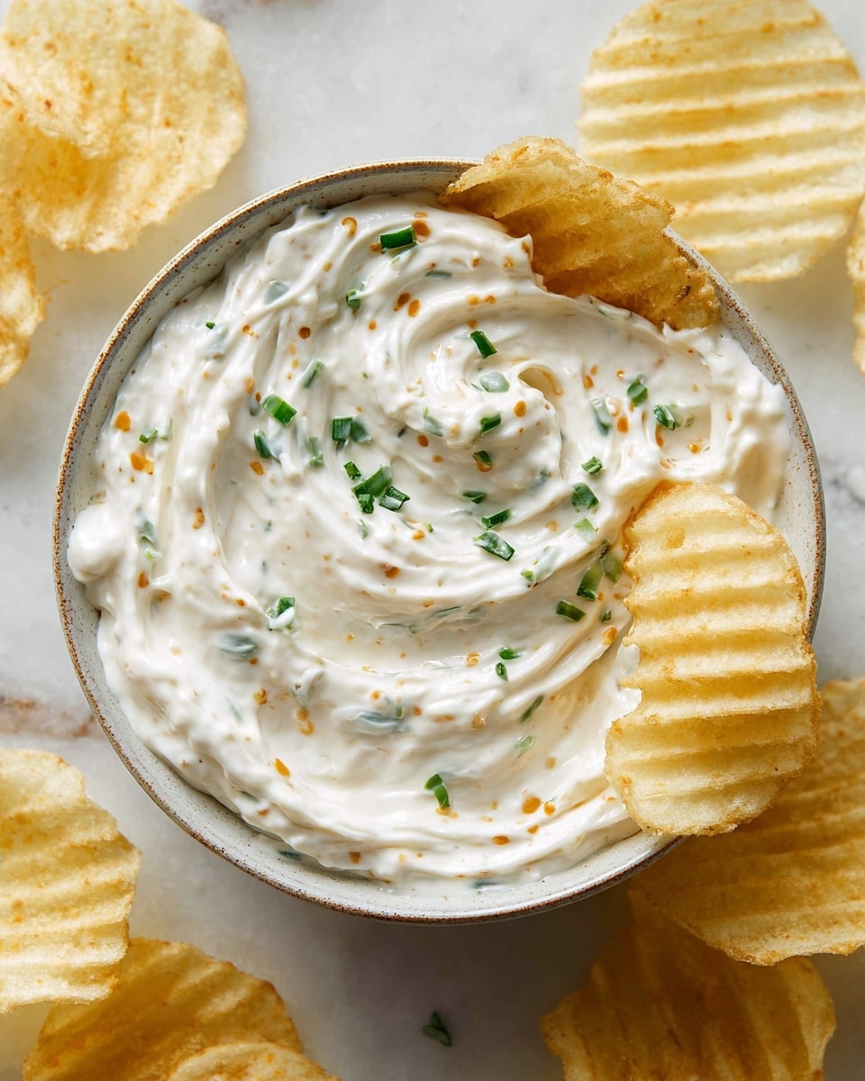 A close-up view of a white ceramic bowl filled with thick, creamy white dip with small green chive pieces and tiny orange flakes mixed evenly throughout. A single rippled yellow potato chip is dipped into the smooth swirl of the dip on the right side of the bowl. The bowl sits on a white marbled surface scattered with several crinkled potato chips in light yellow tones. photo taken with an iphone --ar 4:5 --v 7