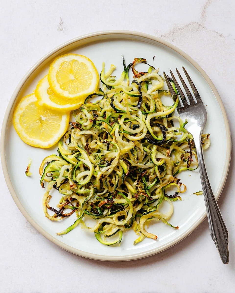 On a white plate, there is a single layer of crispy cooked spiralized zucchini noodles with a mix of light and dark green colors, some edges browned from cooking, showing a slightly crunchy texture. On the left side of the plate, two thin lemon slices with bright yellow rinds and translucent juicy centers are placed side by side. A silver fork rests on the right side of the plate with its prongs facing inward. The plate is set on a white marbled surface. photo taken with an iphone --ar 4:5 --v 7