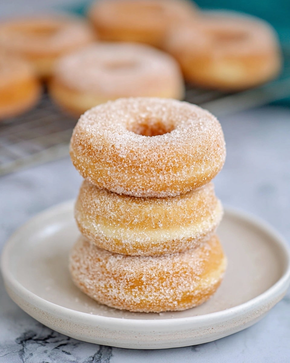 A stack of three sugar-coated donuts is placed in the center of a round white plate with a smooth surface, each donut showing a light golden-brown color covered evenly with granulated sugar, giving a slightly grainy texture. The donuts are thick and fluffy with a small hole in the middle, stacked neatly on top of each other. The background has a soft focus, showing more donuts resting on a cooling rack, while the plate sits on a white marbled texture surface. photo taken with an iphone --ar 4:5 --v 7