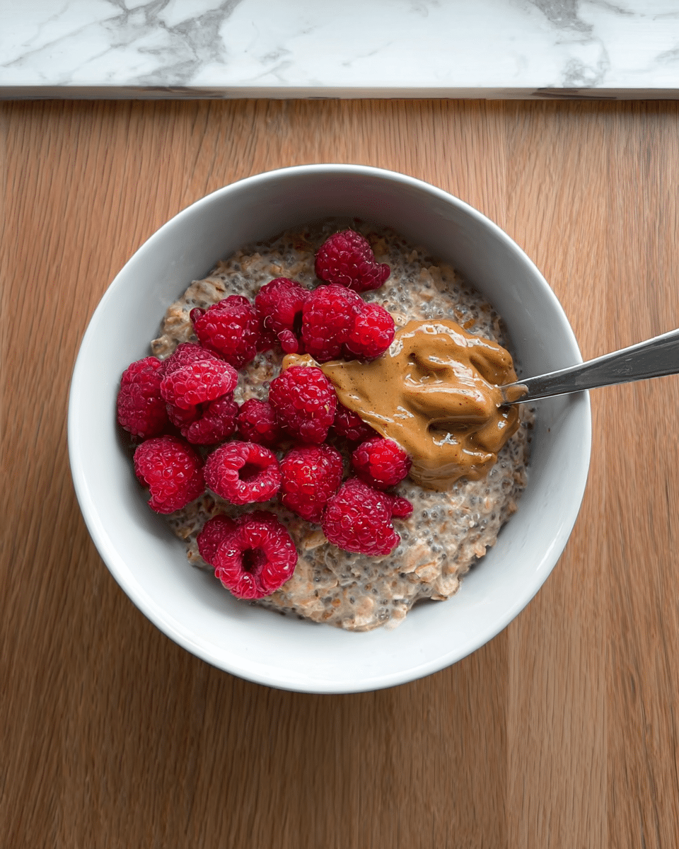 A white bowl filled with creamy overnight oats mixed with chia seeds, showing a thick and somewhat grainy texture as the base layer. On top, a golden-brown dollop of peanut butter spread partially lifting with a silver spoon resting inside the bowl. Bright red fresh raspberries are scattered across the top, adding a vibrant color contrast to the neutral oats and peanut butter. The bowl sits on a wooden surface with a white marbled texture background visible above. Photo taken with an iphone --ar 4:5 --v 7