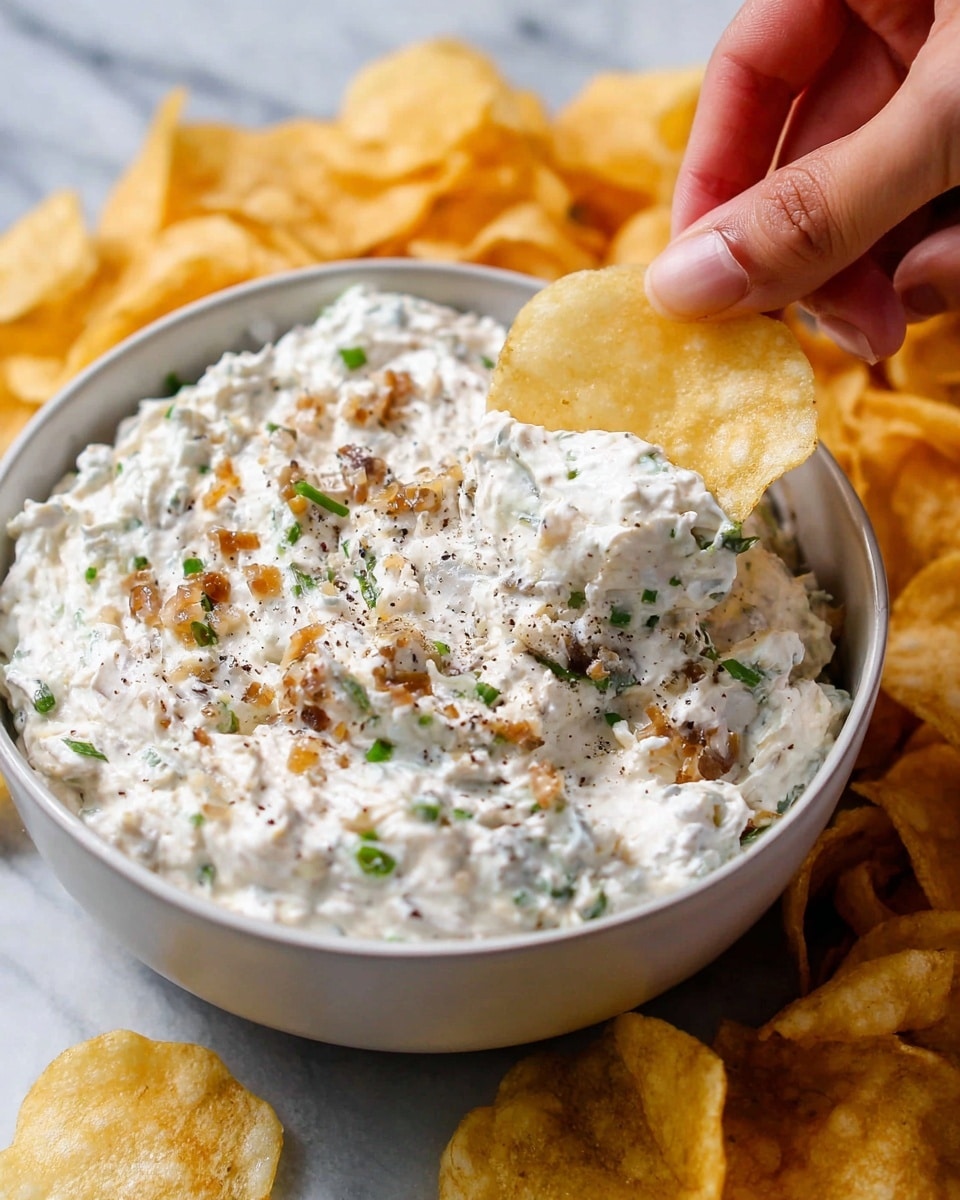 A close-up of a thick creamy dip with visible chopped green herbs and small cooked golden onion pieces mixed in, sitting in a white bowl. The dip is white with specks of black pepper and green herbs spread evenly throughout, giving it a textured and chunky look. Around the bowl, there are pale yellow potato chips scattered, with one chip being held by a woman's hand dipping into the creamy mixture. The background shows a white marbled texture surface. photo taken with an iphone --ar 4:5 --v 7