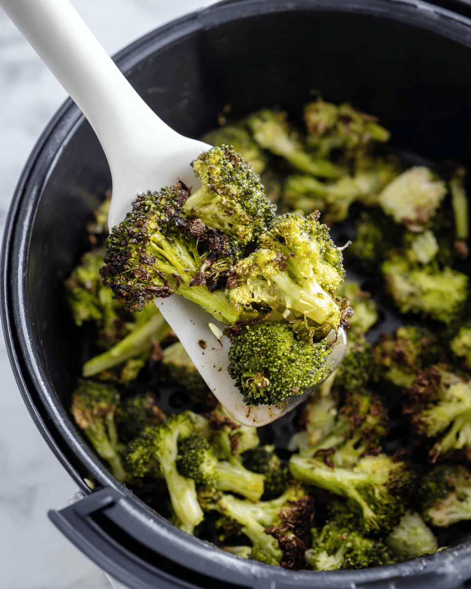 The image shows a white spatula lifting roasted broccoli pieces from a black air fryer basket. The broccoli is lightly browned with some crisp, darkened edges and vibrant green stems and florets, showing a mix of textures from tender to crunchy. The broccoli fills the basket with small uneven pieces, and the white spatula is positioned diagonally, holding a generous portion in the center. The background has a white marbled texture, highlighting the cooking scene. photo taken with an iphone --ar 4:5 --v 7