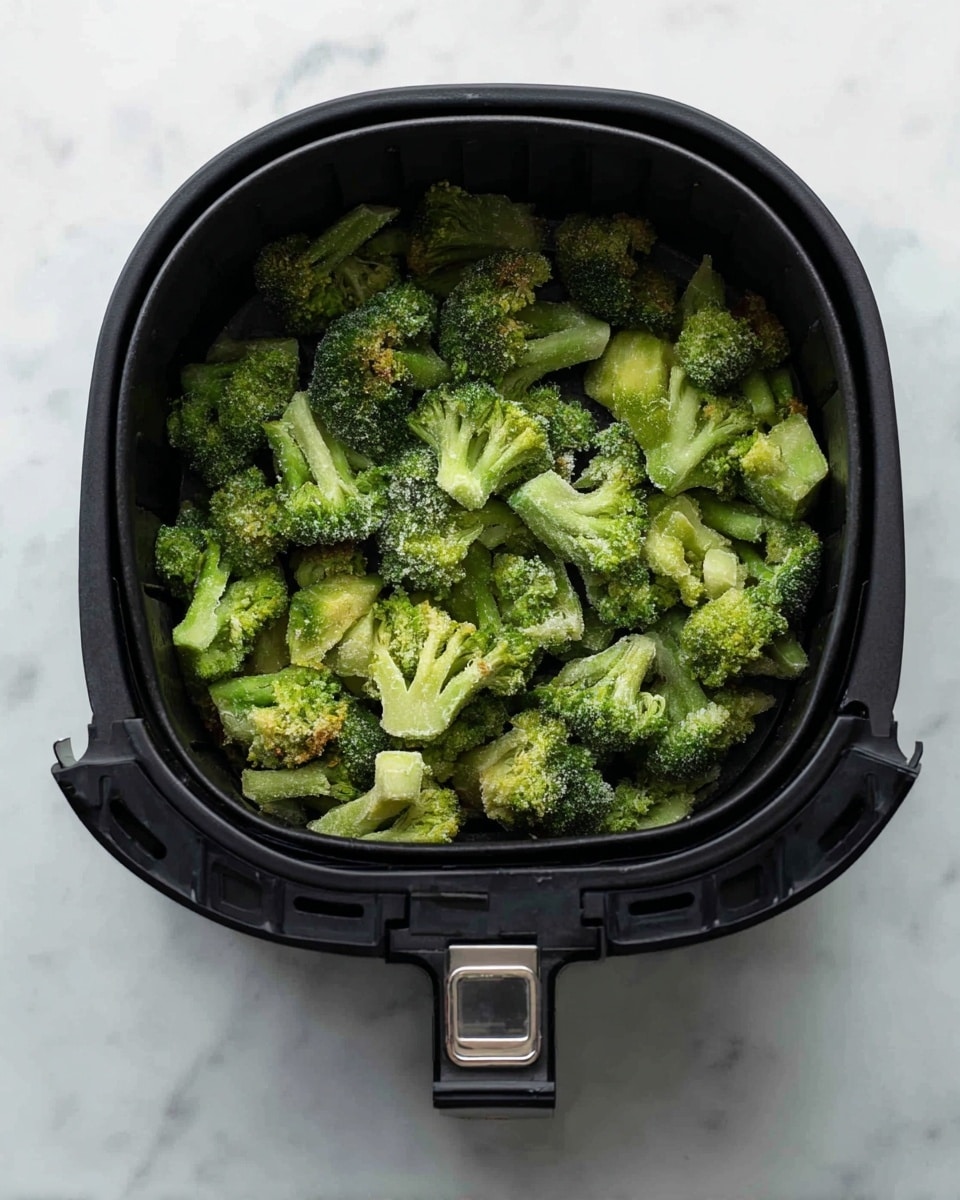 A black air fryer basket filled with bright green broccoli florets that have a slightly frosty texture, showing they are frozen. The broccoli pieces are uneven in size and shape, covering the entire basket surface. The basket has a shiny silver handle, and it is placed on a white marbled surface. photo taken with an iphone --ar 4:5 --v 7