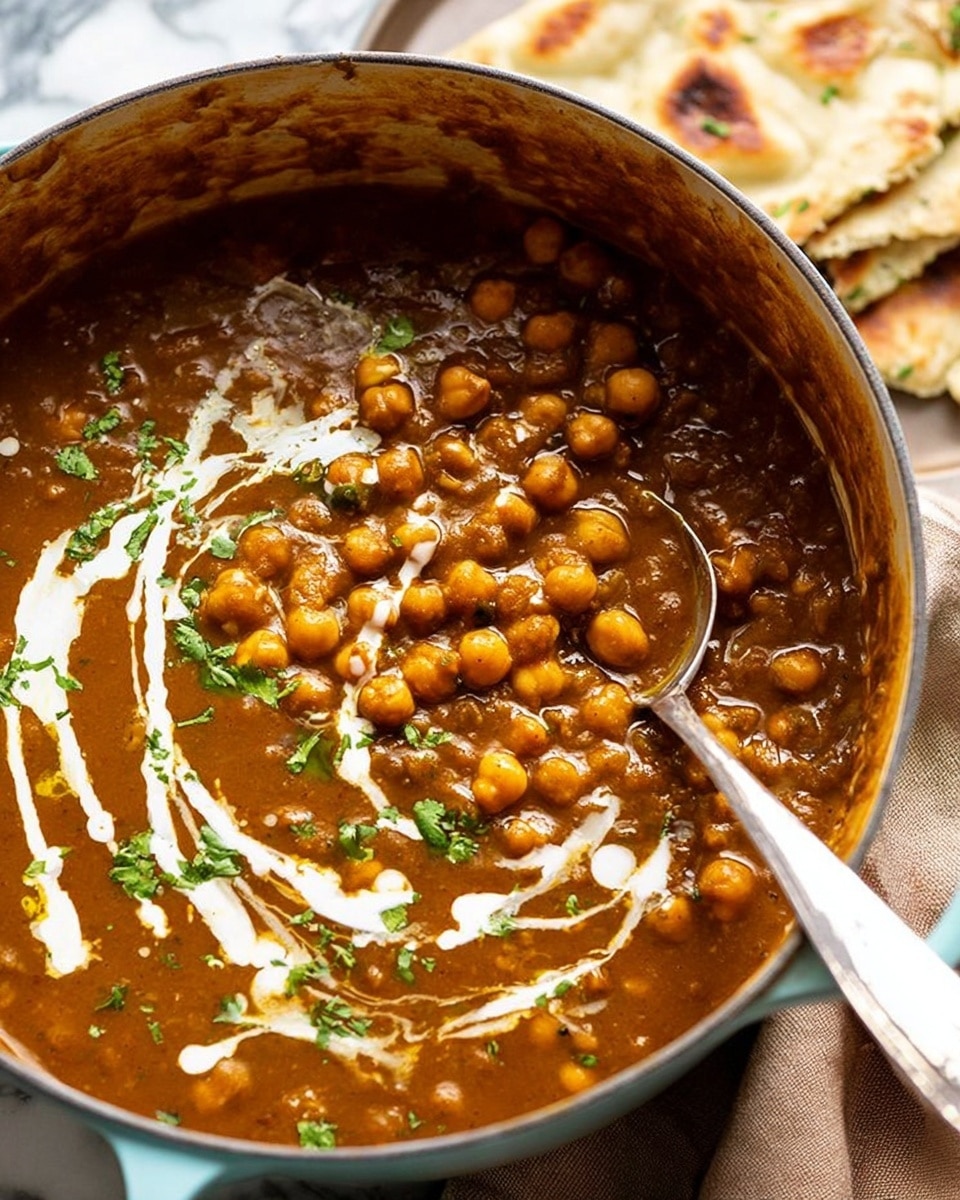A close-up view of a large pot filled with a thick, brown chickpea stew, showing whole chickpeas floating in a rich, slightly chunky sauce. The stew is garnished with swirls of white cream and small pieces of chopped green herbs on top. A metal spoon rests inside the pot, partially submerged in the stew. In the background, there are pieces of flatbread with a light brown color resting on the white marbled surface. The pot has a light blue-gray rim and is placed on a beige cloth. photo taken with an iphone --ar 4:5 --v 7