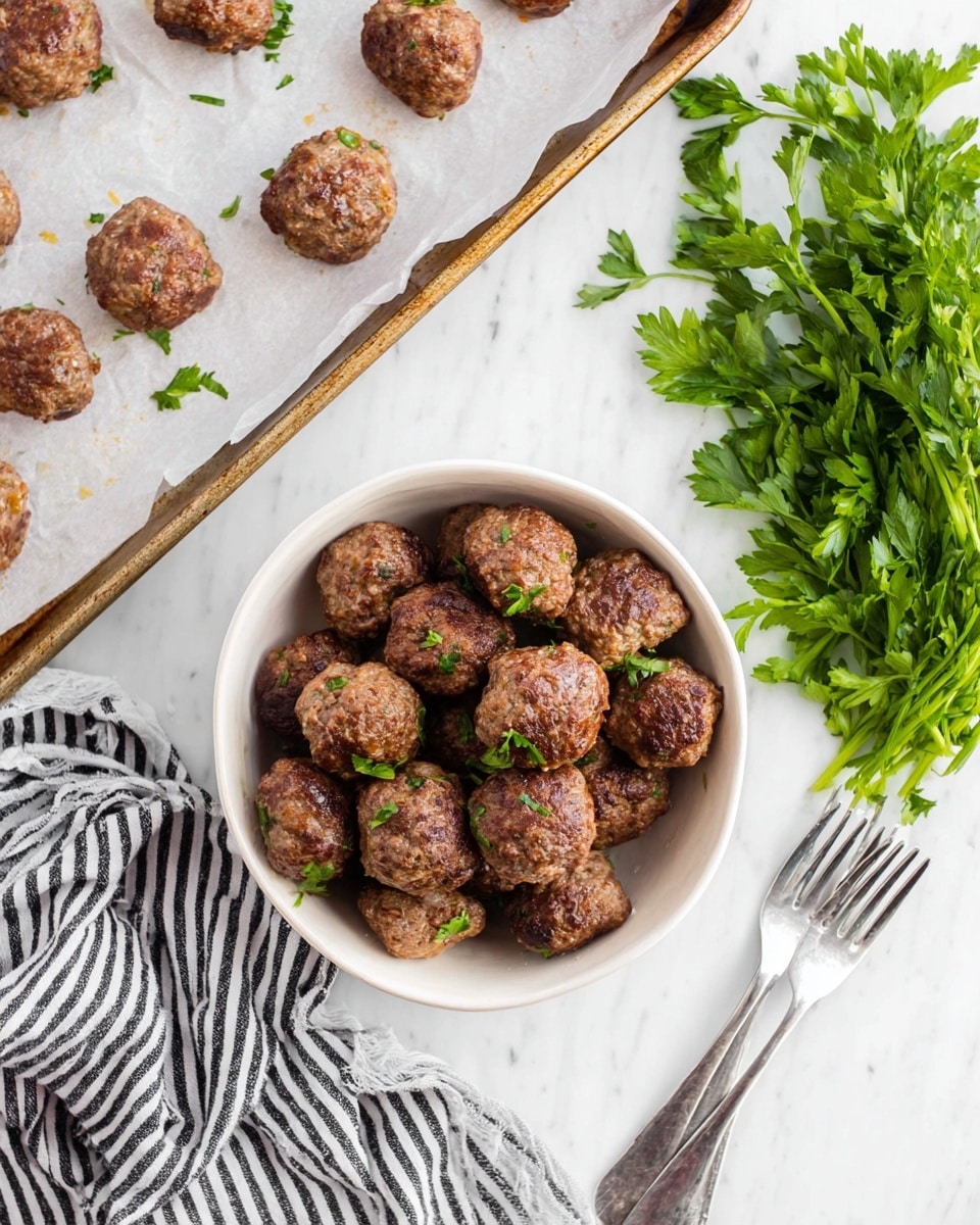 A white bowl is filled with two layers of browned meatballs, each showing a rough, cooked texture with small bits of onion visible. The top meatballs have small green parsley leaves sprinkled on them. Above the bowl, a baking tray with a white parchment paper has a single layer of similar browned meatballs spaced apart. To the right of the bowl, a bunch of fresh green parsley lies on a white marbled surface next to two silver forks. A black and white striped cloth is placed under the bowl on the left side. Photo taken with an iphone --ar 4:5 --v 7